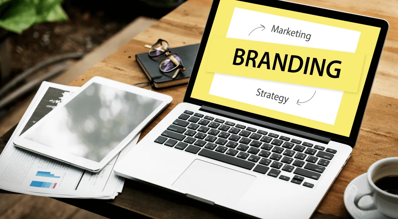 A laptop open on a wooden desk displaying a slide about 'branding' in marketing, accompanied by a tablet, a notebook, glasses, and a cup of coffee.