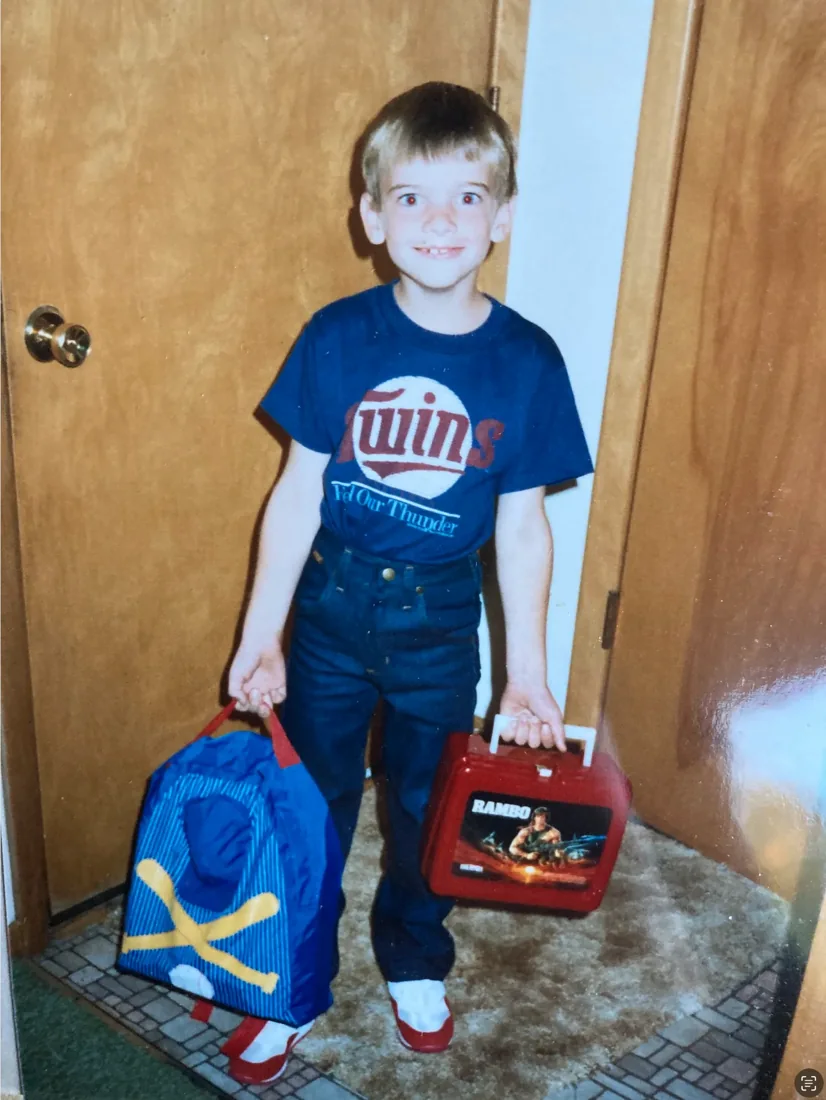 A childhood photo of a boy in a Minnesota Twins shirt with a school bag and lunchbox